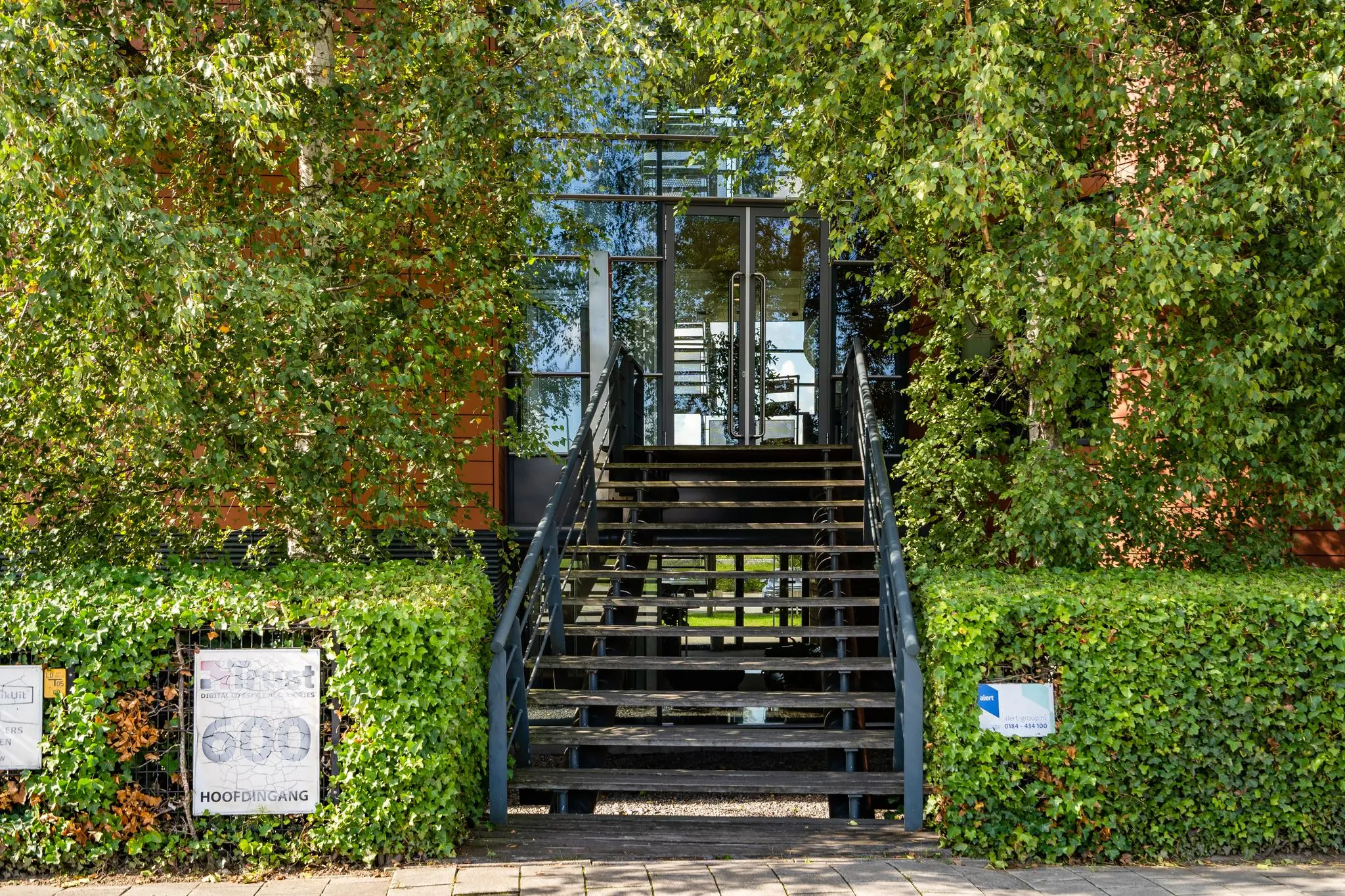 Hoofdingang van het kantoorpand aan de Laan van Barcelona, omgeven door groene beplanting met een trap naar de glazen entree.